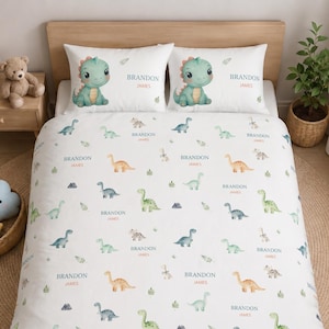 May include: Dinosaur-themed bedding set in white, featuring two pillowcases with a cartoon dinosaur and the name "BRANDON JAMES". The duvet cover has a repeating pattern of dinosaurs, leaves, and the name "BRANDON JAMES".