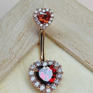 May include: A gold-coloured belly button ring featuring heart-shaped gemstones. The top and bottom hearts are red and encircled by a halo of clear, round gemstones. The jewellery is set against a light wooden background.