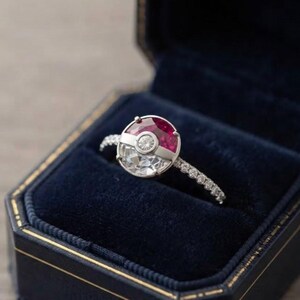 May include: A silver ring featuring a circular design split into a red gemstone top and a clear gemstone bottom, separated by a central silver band with a small round diamond. The ring band is thin and set with a row of small clear stones.