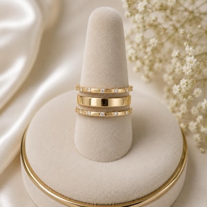 May include: A set of three gold rings stacked together. The top and bottom rings are thin bands featuring small, clear, round-cut stones set at regular intervals. The middle ring is a wider, smooth, polished gold band with no stones.