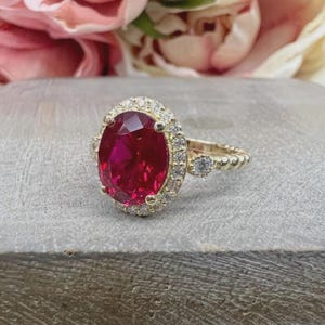 May include: A gold ring with a large, oval-cut ruby gemstone encircled by a halo of smaller, clear gemstones. The ring's band has a textured design. The background features soft pink and white flowers.