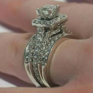Könnte beinhalten: Ein silberfarbener Ring mit einem großen, quadratisch geschliffenen Mittelstein, der von einem Kranz aus kleineren runden Steinen umgeben ist. Die Ringschiene ist breit und in drei Reihen unterteilt, die mit vielen kleinen, funkelnden Steinen besetzt sind.