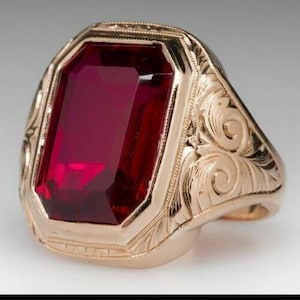 Könnte beinhalten: Ein goldener Ring mit einem großen, rechteckigen, tiefroten Edelstein. Der Ring hat ein verziertes, graviertes Design um den Edelstein und auf dem Band. Der Edelstein ist der Blickfang und fängt das Licht ein.