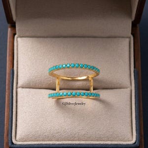 May include: A gold double-band ring featuring two parallel rows of small, round, bright turquoise stones. The ring has a bridge connecting the two bands on each side. The text GJSilverJewelry is printed in black on the beige velvet surface.