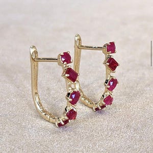 Puede incluir: Un par de pendientes de aro dorados con una serie de gemas rojas vibrantes. Las gemas están engastadas a lo largo de la curva de los aros, creando un diseño brillante y elegante. Los pendientes se muestran sobre un fondo neutro.