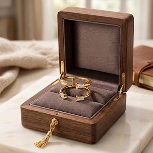 Peut inclure: Une bague ouverte dorée ornée de deux pierres claires taille baguette, placée dans une boîte à bijoux en bois brun foncé avec un intérieur en velours gris doux et un petit pompon doré fixé au mécanisme de fermeture avant.