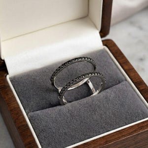 May include: Two thin silver-toned rings featuring a pavé setting of small, round, black stones along the outer band. The rings are positioned upright within a dark wooden jewellery box lined with soft, grey velvet fabric.