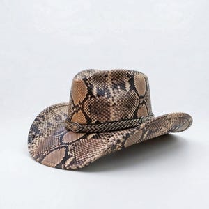 Könnte beinhalten: Ein Cowboyhut mit Schlangenmuster in Braun-, Beige- und Schwarztönen. Der Hut hat ein dekoratives Band um den Kopf und eine breite Krempe.