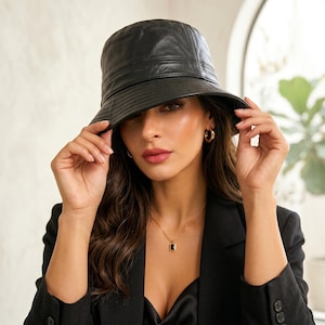 May include: A black bucket hat with a smooth finish and stitched detailing along the brim. The hat features a structured crown and a downward-sloping brim, providing a clean and uniform appearance. The material has a subtle sheen, reflecting light.