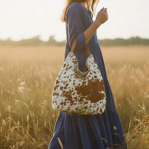 May include: A large tote bag with a cowhide pattern of brown and white spots. The bag has a curved top and brown leather straps. The person in the image is wearing a blue dress and brown boots, walking through a field of tall grass.