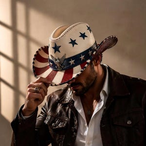 Puede incluir: Un sombrero de vaquero con un diseño patriótico. El sombrero presenta una corona blanca con estrellas azules, un ala a rayas rojas y blancas y una banda azul con estrellas. Un emblema de águila está centrado en la banda. El sombrero se usa con una chaqueta de cuero marrón.