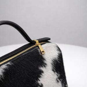 May include: A black and white cowhide handbag with a gold zipper and a black handle. The bag has a rectangular shape and a gold zipper pull. The cowhide pattern is a mix of black and white patches.