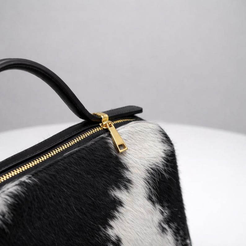 May include: A black and white cowhide handbag with a gold zipper and a black handle. The bag has a rectangular shape and a gold zipper pull. The cowhide pattern is a mix of black and white patches.