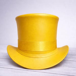 May include: A bright yellow top hat with a wide brim and a band around the crown. The hat is made of a smooth material and sits on a light-colored wooden surface. The hat's design is classic and elegant.