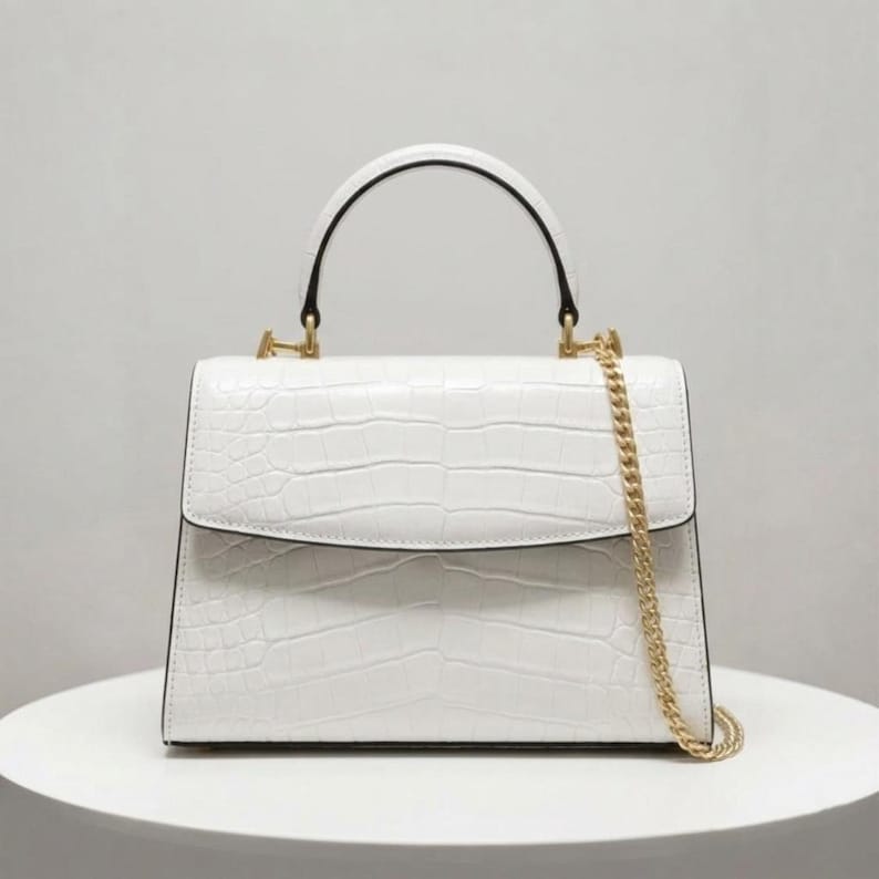 May include: White handbag with a crocodile texture, featuring a top handle and a gold chain strap. The bag has a rectangular shape with a flap closure. The handle is black and curved.
