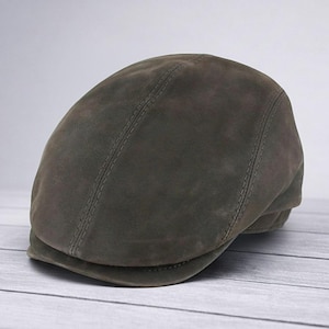 Puede incluir: Gorra plana verde oliva oscuro hecha de un material similar al ante. La gorra tiene una corona redondeada y una visera pequeña y rígida. La parte superior de la gorra presenta una costura central. La gorra está sobre una superficie de madera clara.