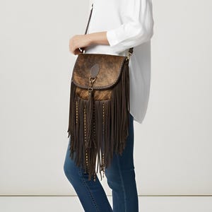 May include: A brown leather crossbody bag with a flap closure and long fringe. The bag features a cowhide front panel with a natural pattern. Gold-colored studs accent the fringe.