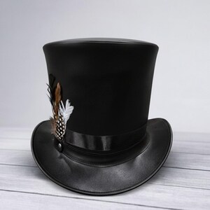 Puede incluir: Sombrero de copa negro con ala ancha y un adorno de plumas decorativas. El sombrero presenta una cinta de raso y un detalle de bot&oacute;n. Las plumas son marrones, blancas y negras.