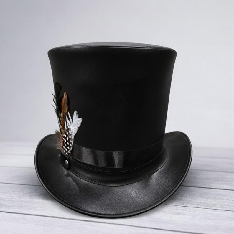 Puede incluir: Sombrero de copa negro con ala ancha y un adorno de plumas decorativas. El sombrero presenta una cinta de raso y un detalle de bot&oacute;n. Las plumas son marrones, blancas y negras.