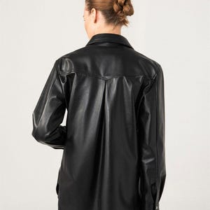 May include: Black long-sleeve button-down shirt made of faux leather. The shirt has a classic collar, buttoned cuffs, and a slightly oversized fit. The back features a vertical pleat detail.