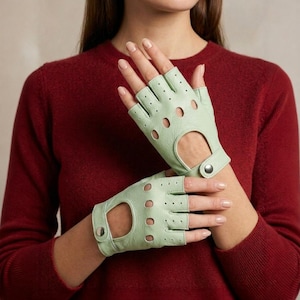 Könnte beinhalten: Hellgrüne fingerlose Handschuhe mit kreisförmigen Aussparungen und einem Handgelenkriemen mit silbernem Druckknopfverschluss. Die Handschuhe sind aus weichem Material und haben ein klassisches Design.