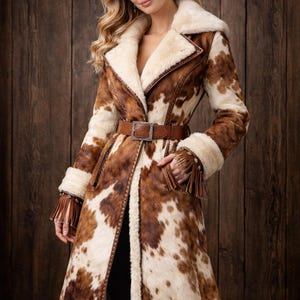 May include: A long, cowhide-patterned coat with a cream-colored shearling lining and collar. The coat features a brown leather belt with a silver buckle and fringe detailing on the cuffs. The coat is open in the front, revealing a black outfit.