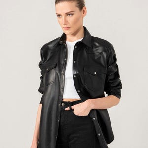May include: Black leather shirt with two chest pockets and button closures. The shirt is paired with black jeans and a white top. The model is wearing the shirt open. The shirt has a classic collar.