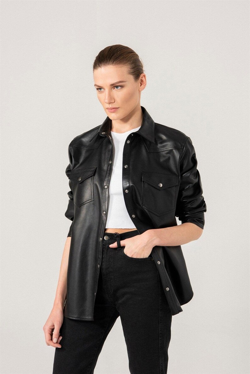 May include: Black leather shirt with two chest pockets and button closures. The shirt is paired with black jeans and a white top. The model is wearing the shirt open. The shirt has a classic collar.