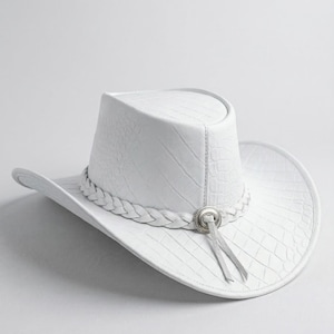 May include: A white cowboy hat with a textured, crocodile-like pattern. The hat features a braided band around the crown and a silver-toned decorative element with a tassel. The brim is curved, and the hat is set against a white background.