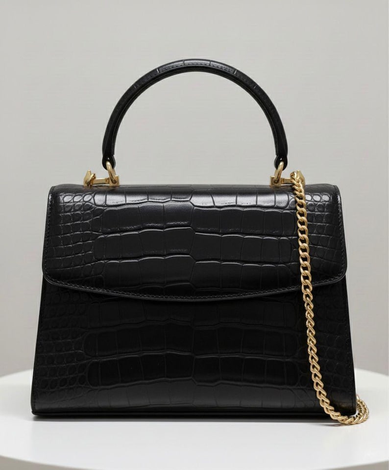 May include: A black handbag with a crocodile-effect texture. It features a top handle and a gold chain strap. The bag has gold-toned hardware and a rectangular shape with a flap closure.