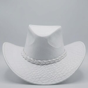 May include: A white cowboy hat with a textured, embossed pattern. The hat features a braided band around the crown and a wide brim. The hat's design includes a crocodile-like texture on the brim and crown.