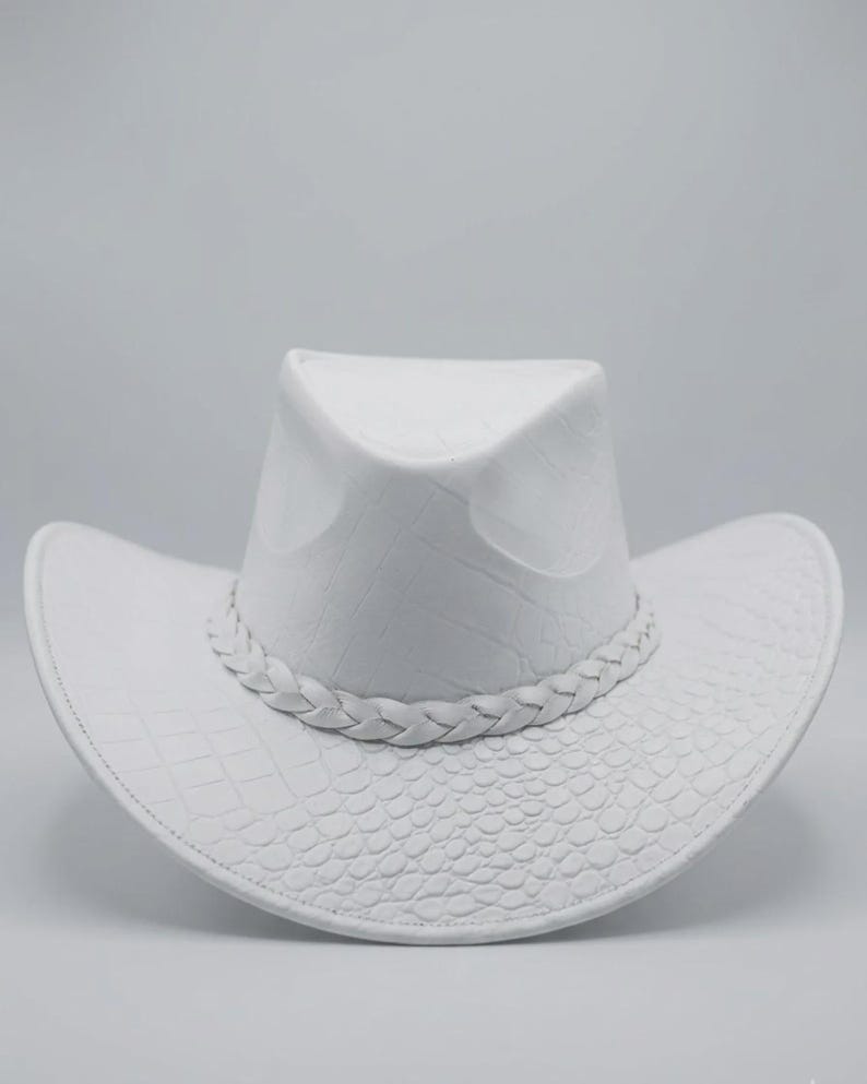 May include: A white cowboy hat with a textured, embossed pattern. The hat features a braided band around the crown and a wide brim. The hat's design includes a crocodile-like texture on the brim and crown.