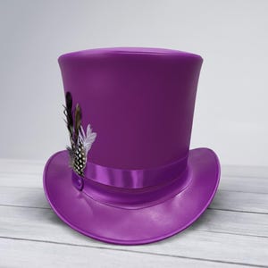 Puede incluir: Un llamativo sombrero de copa morado con una cinta de raso y un adorno de plumas decorativas. El sombrero tiene un ala ancha y una copa alta y cil&iacute;ndrica. Las plumas son negras, marrones y blancas.