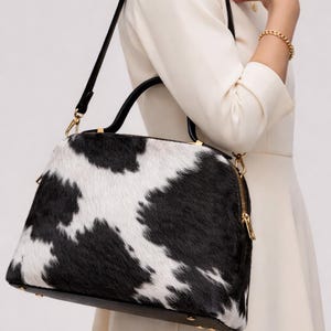 May include: A black and white cowhide handbag with a black handle and gold-colored zipper and hardware. The bag has a rounded shape and a black base. The bag is being carried by a person wearing a white dress.
