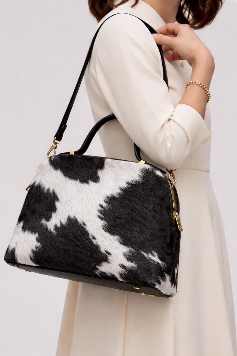 May include: A black and white cowhide handbag with a black handle and gold-colored zipper and hardware. The bag has a rounded shape and a black base. The bag is being carried by a person wearing a white dress.