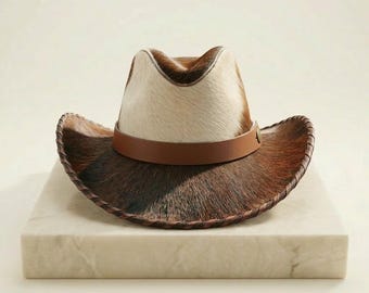 Puede incluir: Un sombrero de vaquero con un patrón de piel de vaca marrón y blanco. El sombrero presenta una banda de cuero marrón y un ala cosida. El sombrero se exhibe sobre un bloque de mármol blanco.