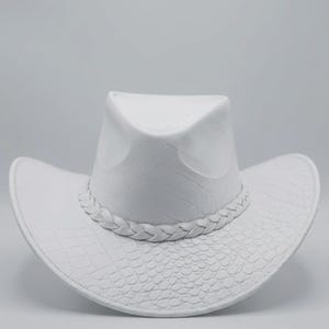 May include: A white cowboy hat with a textured, embossed pattern. The hat features a braided band around the crown and a curved brim. The hat's design includes a crocodile-like texture on the brim and crown.