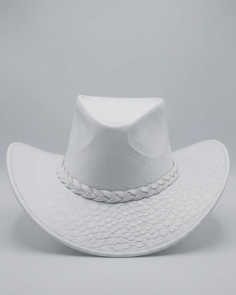 May include: A white cowboy hat with a textured, embossed pattern. The hat features a braided band around the crown and a curved brim. The hat's design includes a crocodile-like texture on the brim and crown.