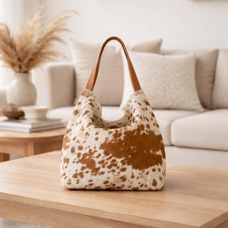 May include: A cowhide-patterned tote bag with brown and white spots. The bag has a curved top and brown leather handles. The bag is sitting on a wooden table in a living room setting.