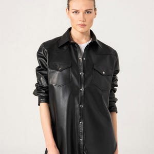 May include: Black leather shirt with two chest pockets and silver snap closures. The long-sleeved shirt has a classic collar and is worn over a white t-shirt. The shirt is a stylish piece of apparel.