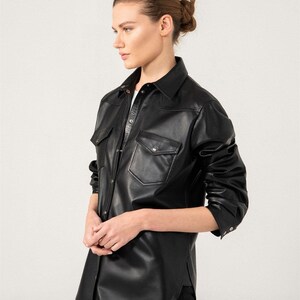 May include: A black leather shirt with two chest pockets and a collared neckline. The long-sleeved shirt has a button-down front and is worn with black pants. The model is facing to the side against a white background.