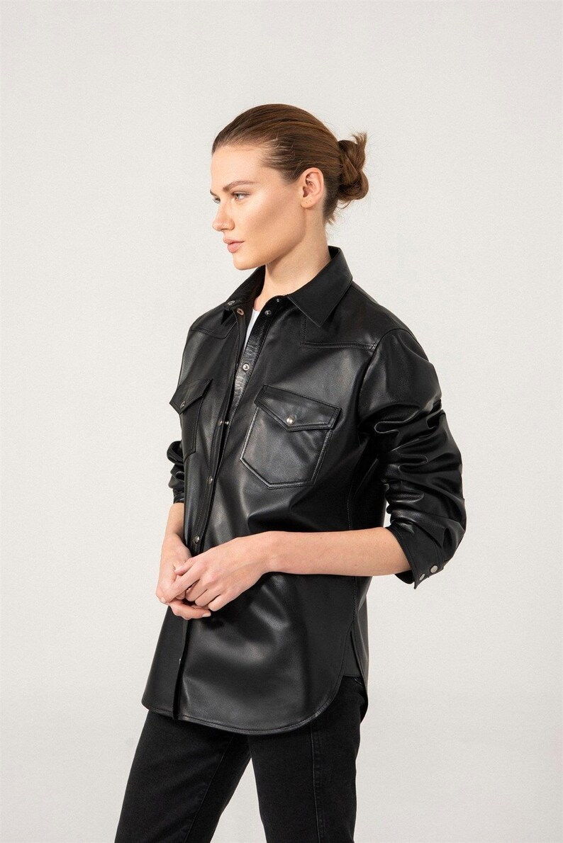 May include: A black leather shirt with two chest pockets and a collared neckline. The long-sleeved shirt has a button-down front and is worn with black pants. The model is facing to the side against a white background.