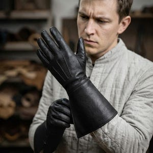 Puede incluir: Guantes de cuero negro con largas guanteletes. Los guantes están hechos de cuero texturizado y tienen detalles de costura visibles. Los guanteletes se extienden por el antebrazo. Los guantes parecen nuevos y sin usar.