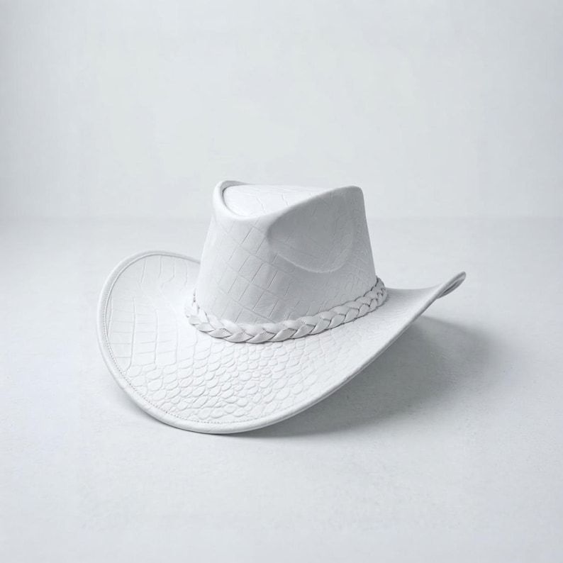 May include: A white cowboy hat with a textured, faux-leather appearance. The hat features a braided band around the crown and a wide brim. The hat's design includes a subtle embossed pattern, adding to its visual appeal.