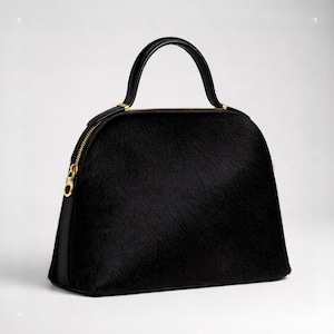 May include: A black handbag with a rounded shape and a top handle. The bag features a textured surface, a gold-colored zipper, and a black leather handle. The bag is set against a white background.