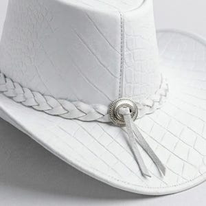 May include: A white cowboy hat with a textured, crocodile-like pattern. The hat features a braided band and a silver-toned decorative element with two hanging leather ties. The hat's brim is curved, and the overall design is clean and stylish.