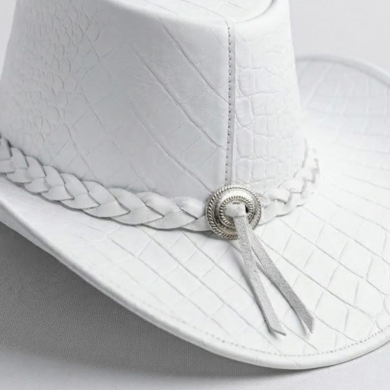 May include: A white cowboy hat with a textured, crocodile-like pattern. The hat features a braided band and a silver-toned decorative element with two hanging leather ties. The hat's brim is curved, and the overall design is clean and stylish.