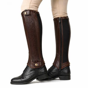 May include: Brown leather riding half chaps with vertical ribbed detailing and gold-tone button closures. The chaps are paired with black leather riding boots. A black elastic panel runs along the back of the chaps. The boots and chaps are shown against a white background.