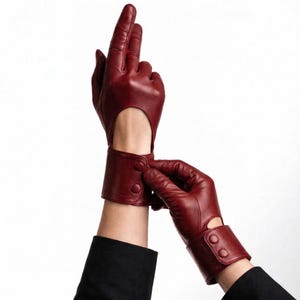 May include: A pair of burgundy leather gloves with a wrist cutout and button closure. The gloves are a deep red color and appear to be made of soft leather. The wrist cuffs have a button closure.