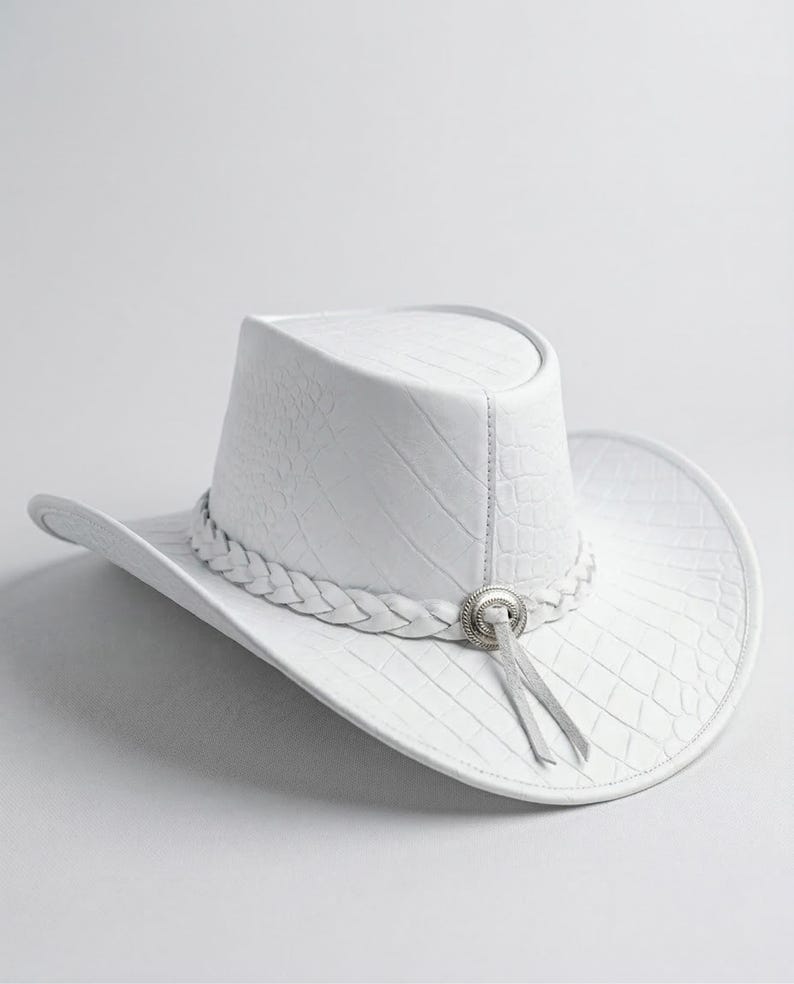 May include: A white cowboy hat with a textured, crocodile-like pattern. The hat features a braided band around the crown and a silver-toned decorative element with a tassel. The brim is curved, and the hat is set against a white background.